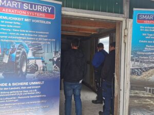 Drei Männer stehen in einem schmalen Raum zwischen zwei großen blauen Bannern mit Informationen zu SMART SLURRY Aeration Systems beim Tag des offenen Güllebehälters bei Beerepoot